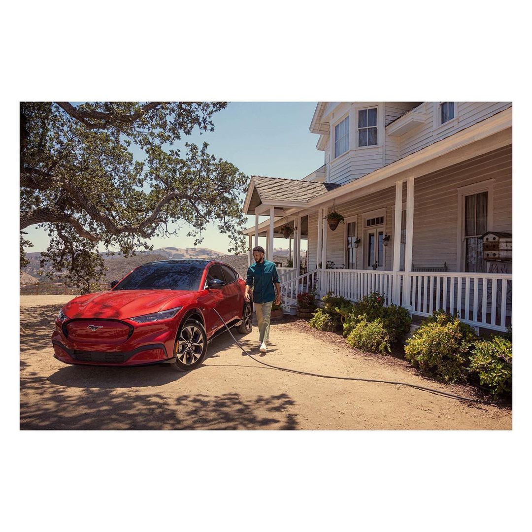 A red Ford Mustang Mach-E is being washed by an adult male in front of a large white two-story home, with a mountain background and blue sky.