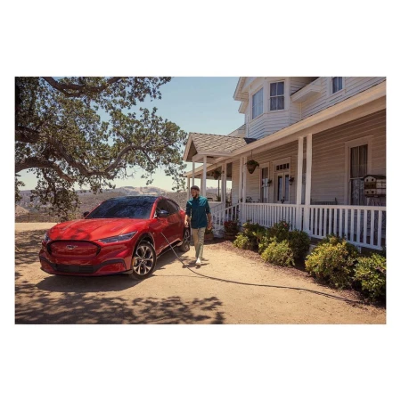 A red Ford Mustang Mach-E is being washed by an adult male in front of a large white two-story home, with a mountain background and blue sky.