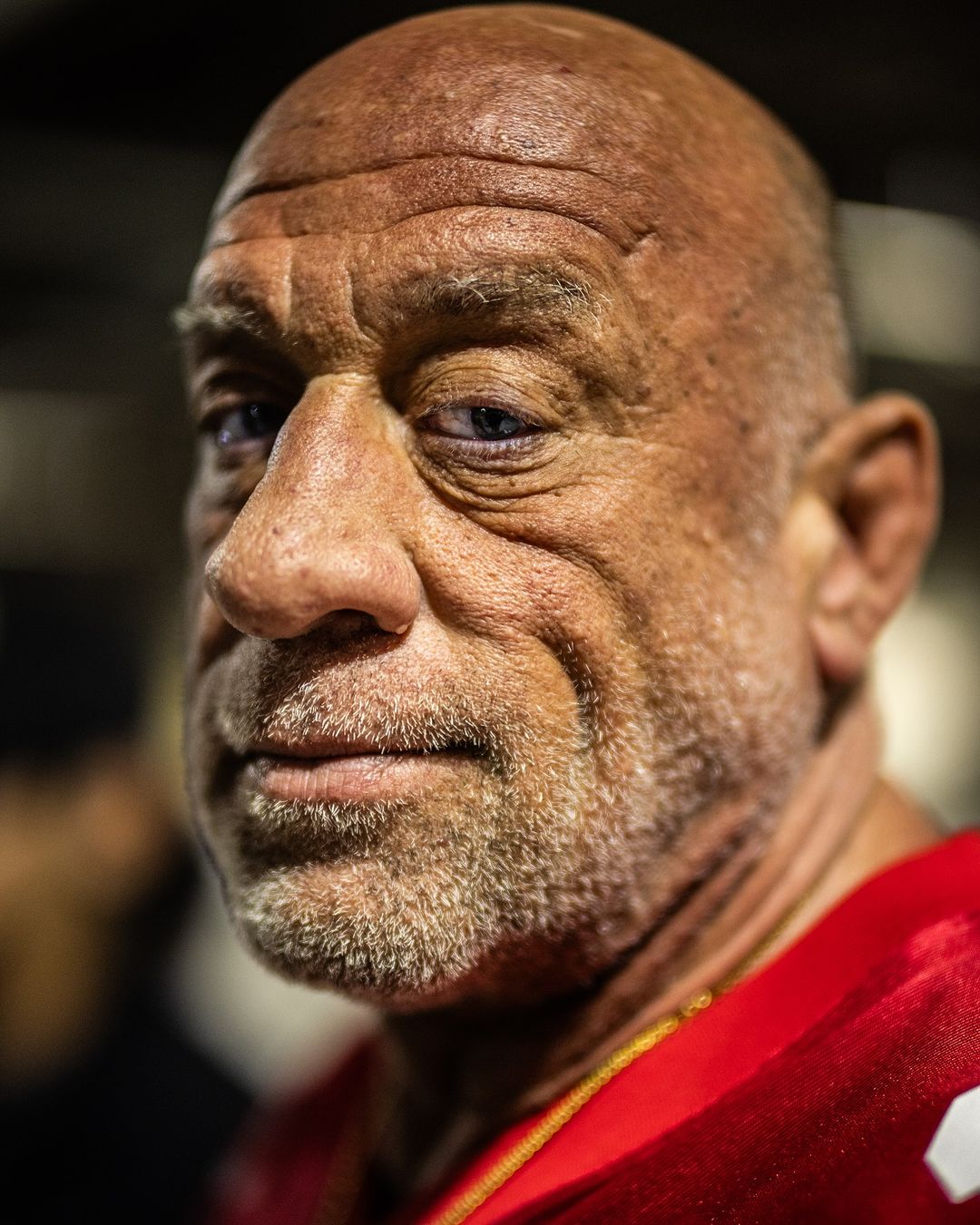 Close-up portrait of a bald adult male with a light beard, wearing a red shirt. The image features a shallow depth of field and dark lighting.