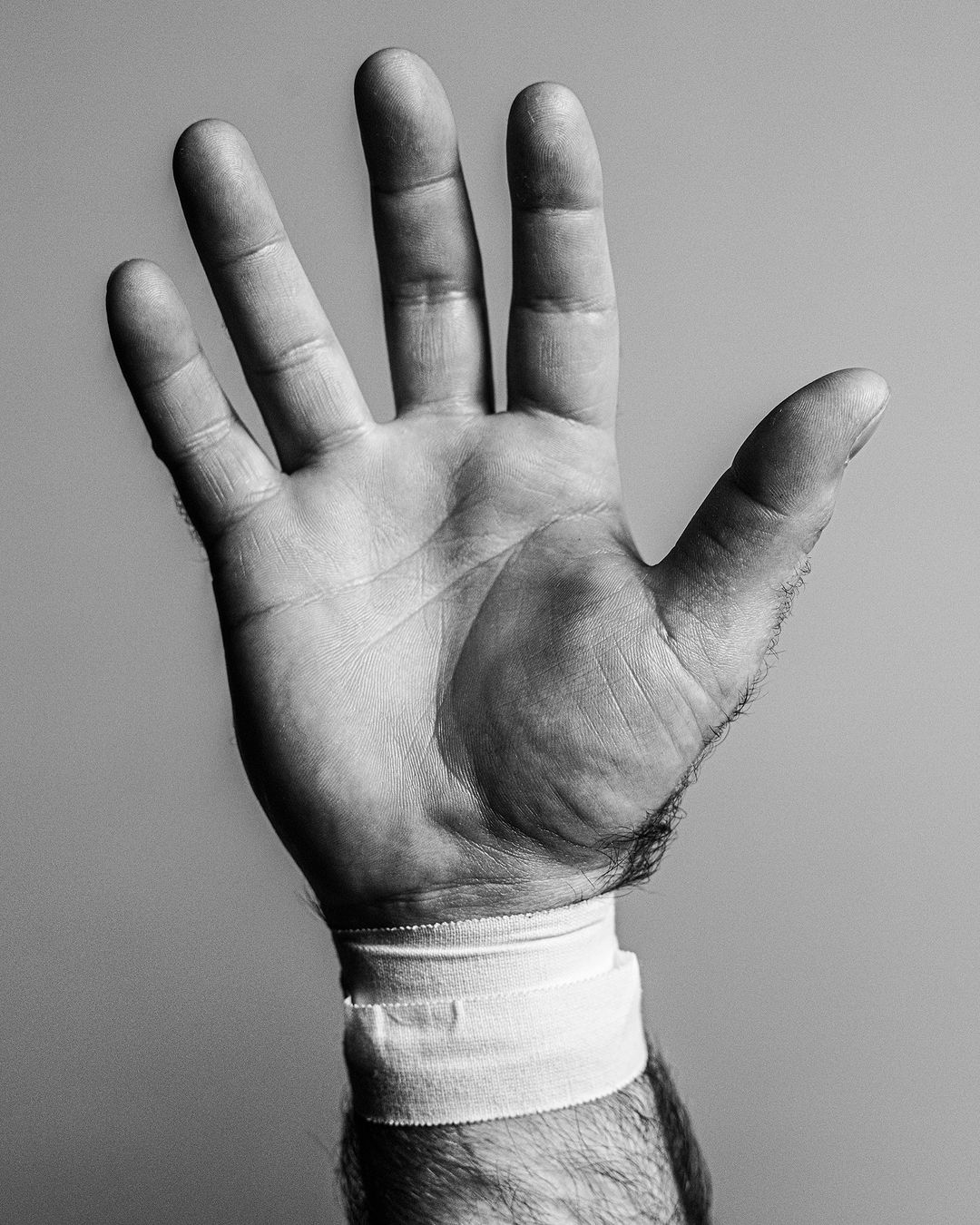 Monochrome image of a person's open hand, fingers spread, with a white bandage around the wrist, set against a grey background.