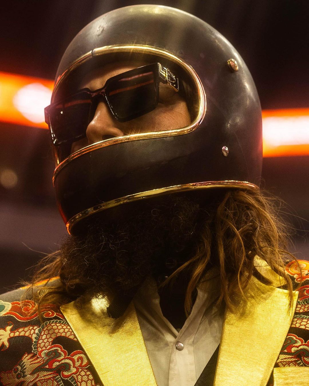 Portrait of a person wearing a black motorcycle helmet with gold trim, dark sunglasses, and an ornate yellow and red jacket.