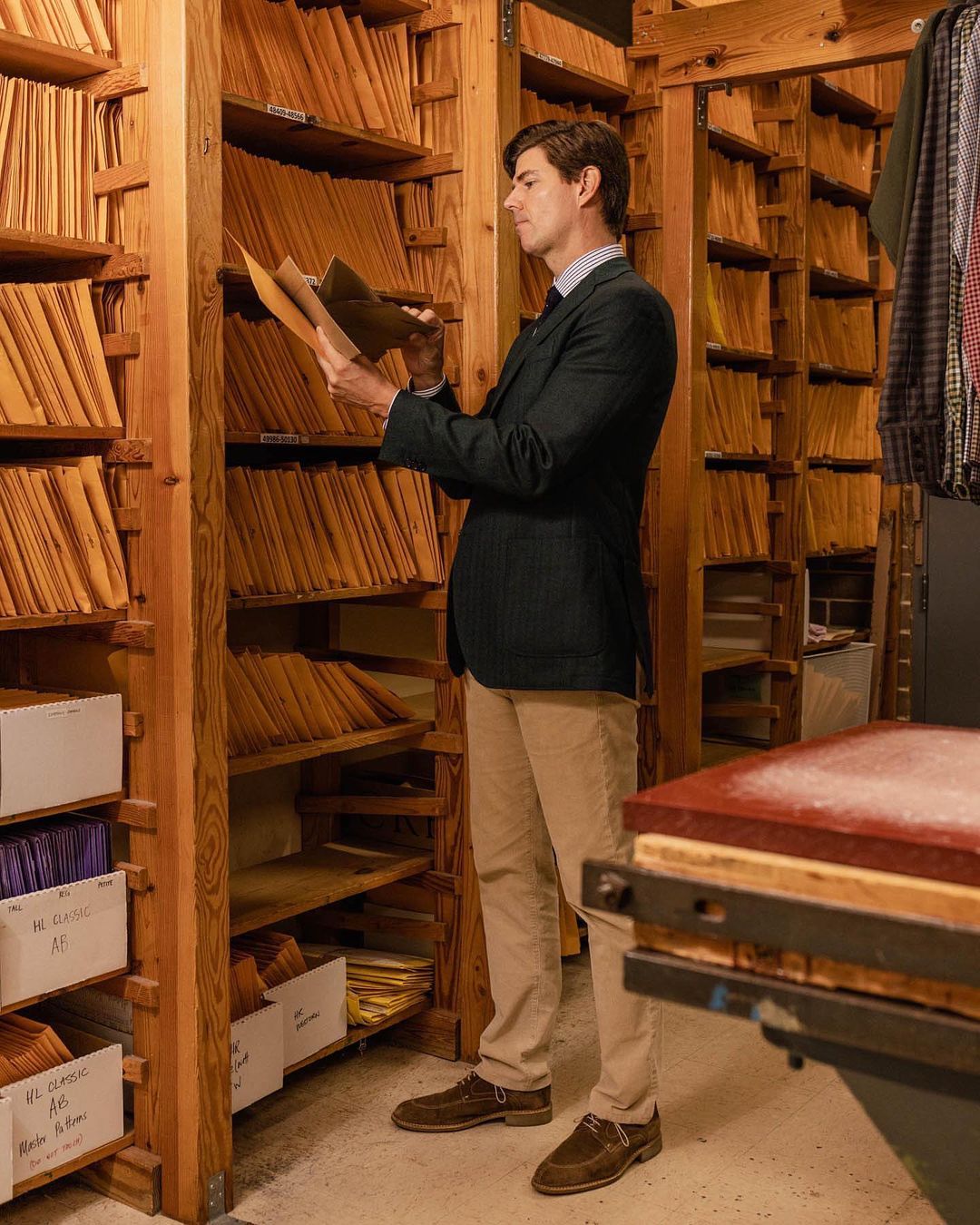 Adult male in a navy blazer stands in a wood-paneled room, reviewing papers on a shelf. His attire is professional, with tan pants and brown shoes.