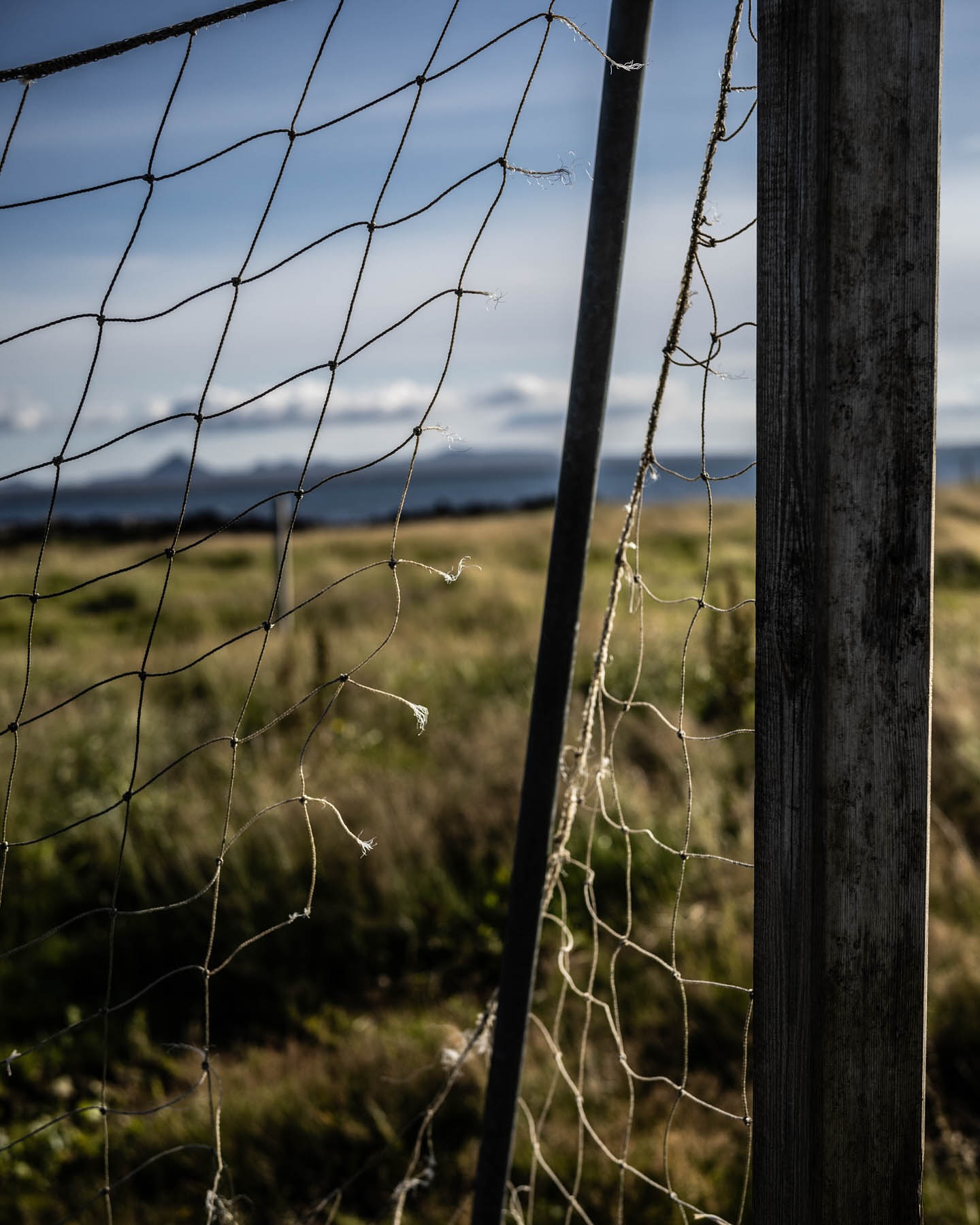 Close-up of a weathered net strung on wooden and metal posts against a background of field and distant hills under a blue sky.