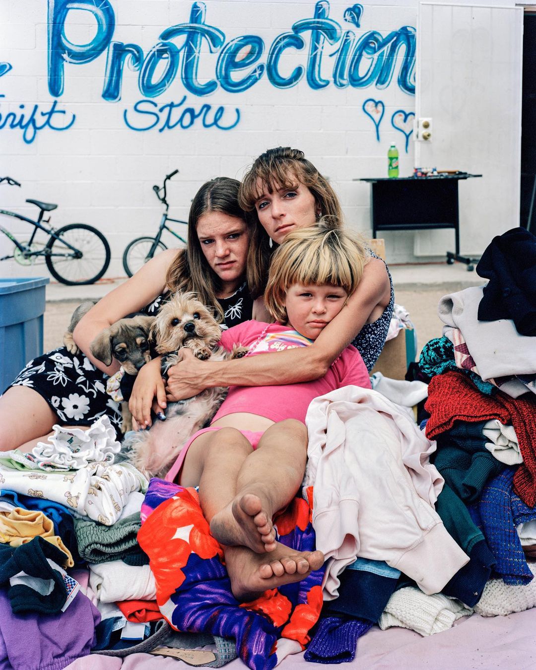 Three people and two dogs pose together, surrounded by clothes, in front of a building marked 'Protection Thrift Store'.