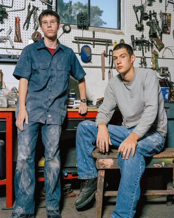 Two adult males are indoors in a workspace, one standing with a blue work shirt and the other sitting on a stool in blue jeans.