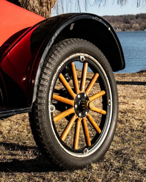 Close-up of vintage red car wheel with wooden spokes parked on grassy ground near calm water and trees under a clear blue sky.