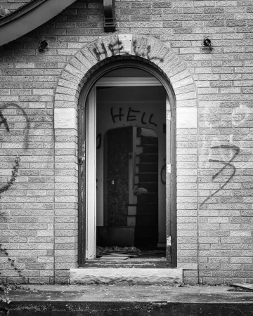 Exterior of an abandoned brick building with a doorway, vandalized with graffiti-style writing, creating a stark and desolate architectural image.