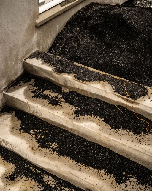 Concrete stairs covered with black granular material lead up to a window, creating an abstract, textured indoor scene.