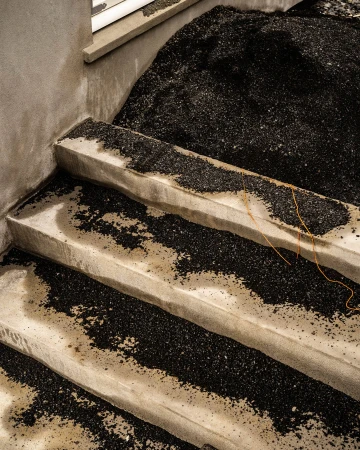 Concrete stairs covered with black granular material lead up to a window, creating an abstract, textured indoor scene.