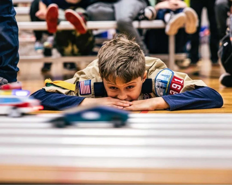 Young boy in Scout uniform lies on floor to watch toy cars race on a track, with spectators and bench in background indoors.