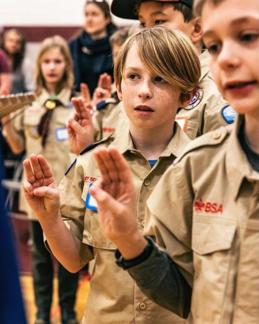 Group of boys in tan scout uniforms raise their right hands, indoors, with other kids and adults in the background.