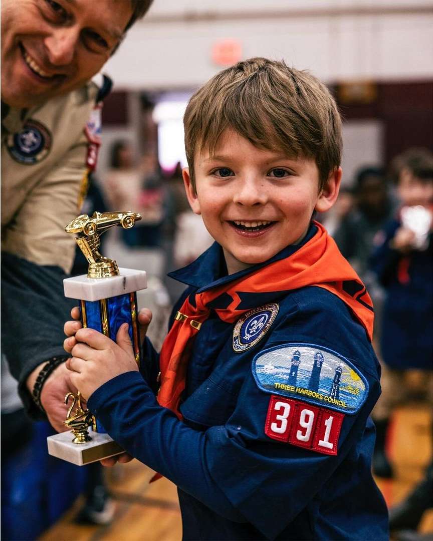 Smiling young boy in uniform holds a trophy with adult in background at indoor event, showcasing achievement and recognition.