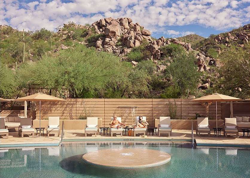 Outdoor swimming pool area with two adults relaxing on lounge chairs, umbrellas, and a desert mountain landscape backdrop.