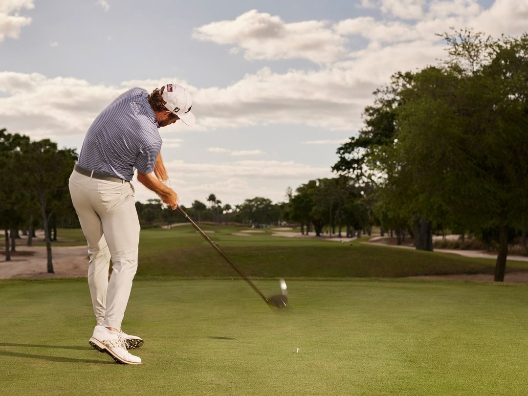 Adult male golfer wearing a cap, light pants, and golf shoes swings a club on a green golf course under a cloudy sky.