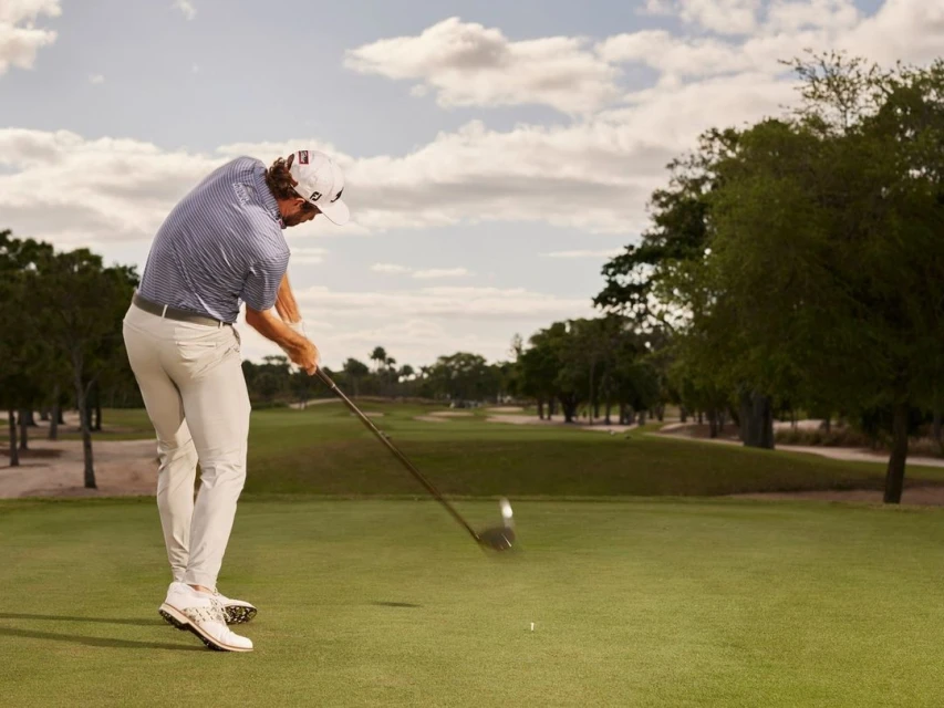 Adult male golfer wearing a cap, light pants, and golf shoes swings a club on a green golf course under a cloudy sky.