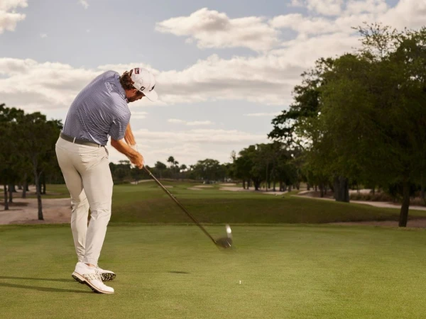 Adult male golfer wearing a cap, light pants, and golf shoes swings a club on a green golf course under a cloudy sky.