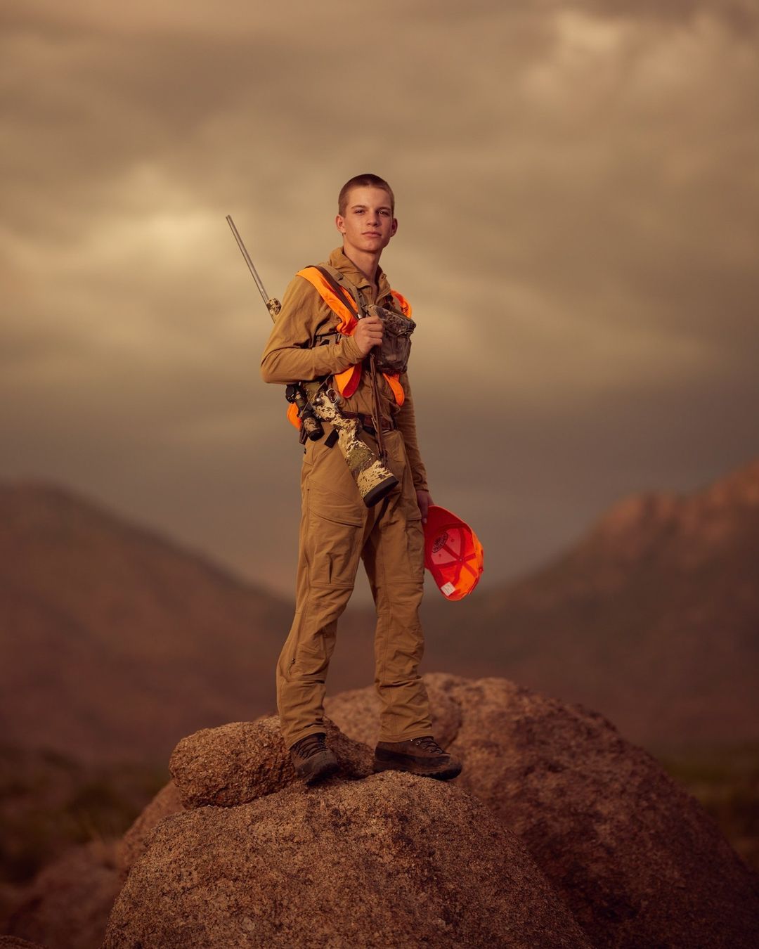 Young hunter in outdoor gear stands on a rock against a mountain backdrop, wearing an orange safety vest and holding a rifle.