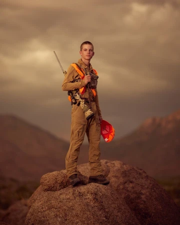 Young hunter in outdoor gear stands on a rock against a mountain backdrop, wearing an orange safety vest and holding a rifle.