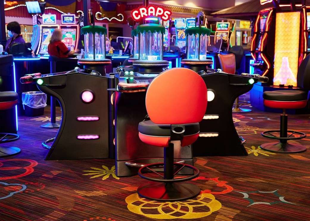 Interior view of a casino featuring craps tables with illuminated signs and chairs, slot machines, and patterned carpeting.