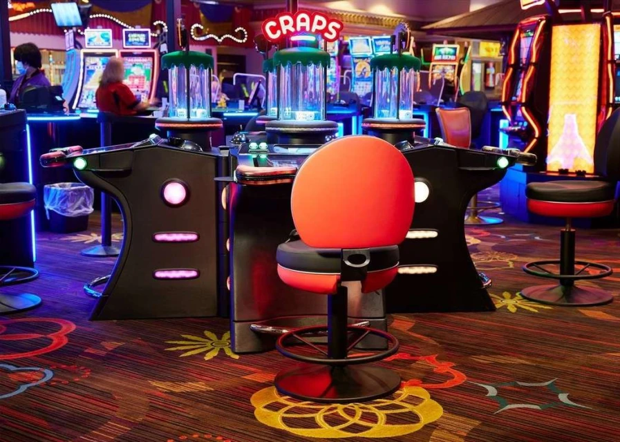 Interior view of a casino featuring craps tables with illuminated signs and chairs, slot machines, and patterned carpeting.