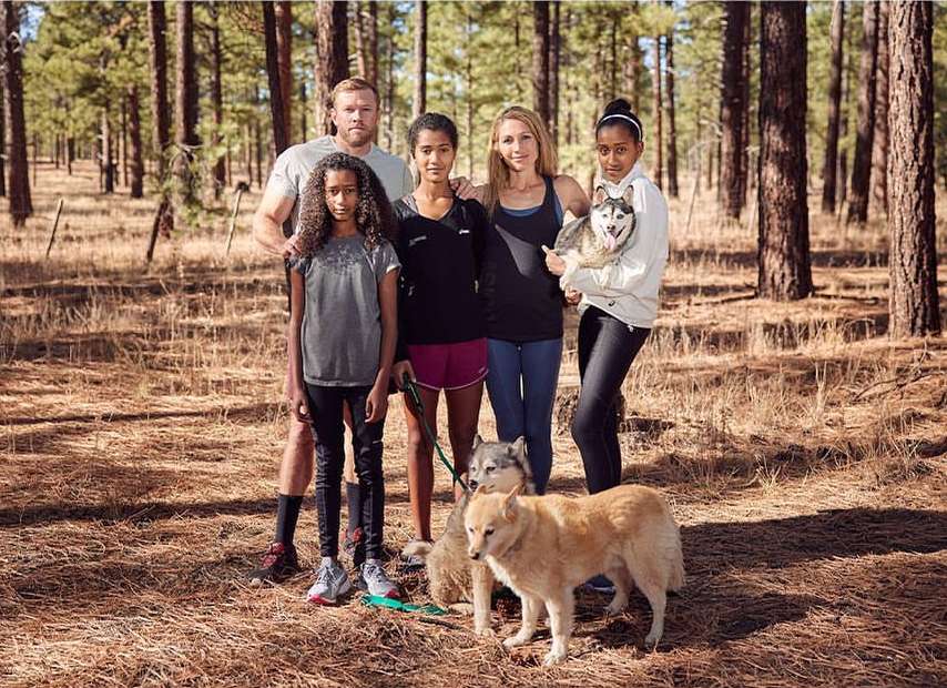 Family of five poses with dogs in a forest. Adults, children, and dogs of different breeds stand together amongst tall trees.