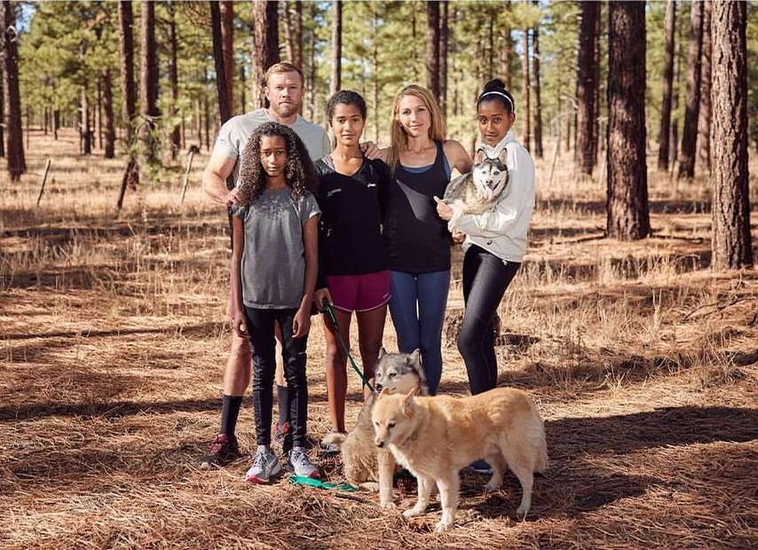 Family of five poses with dogs in a forest. Adults, children, and dogs of different breeds stand together amongst tall trees.