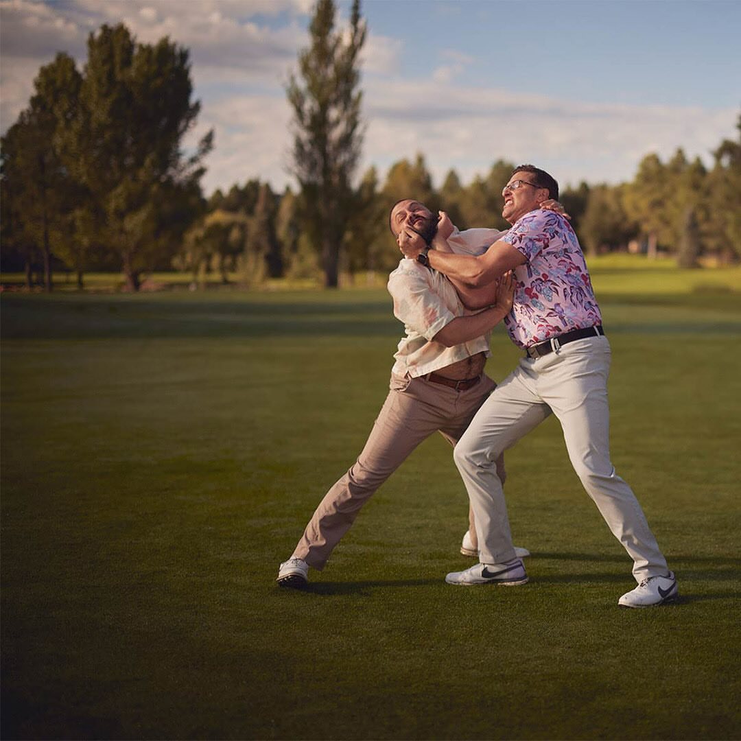 Two adult men in a physical altercation on a golf course, one grabbing the other around the neck, with trees and a cloudy sky in the background.