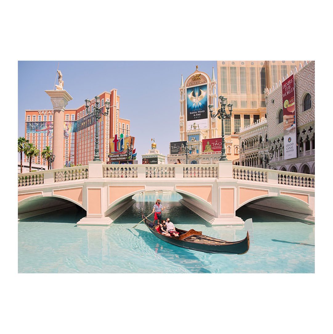 Gondola ride in a canal, tourists being rowed under an arched bridge in a replica Venice setting with Venetian-style buildings.