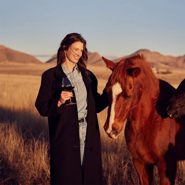 Woman in black coat holds red wine glass next to a brown horse in a grassy field, under a clear sky and distant mountains.