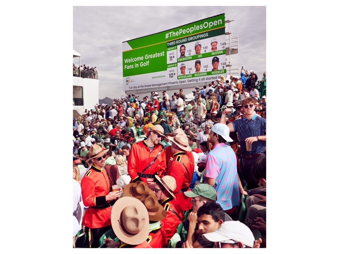 Large crowd of golf fans watches a tournament with a large green and white digital scoreboard displaying golfers and sponsor messages.