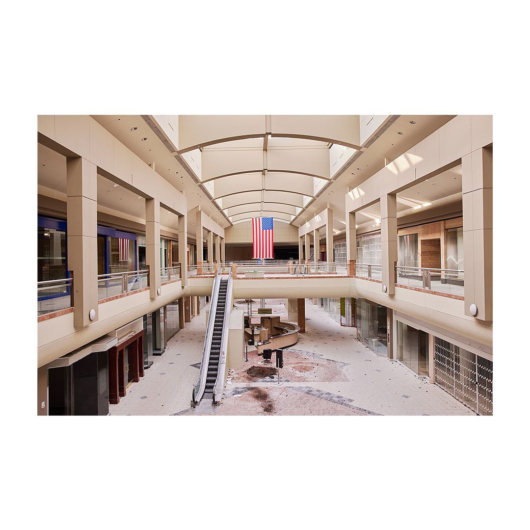 Interior of a mostly-empty two-story shopping mall featuring an escalator, overhead skylights, and a large American flag hanging overhead.