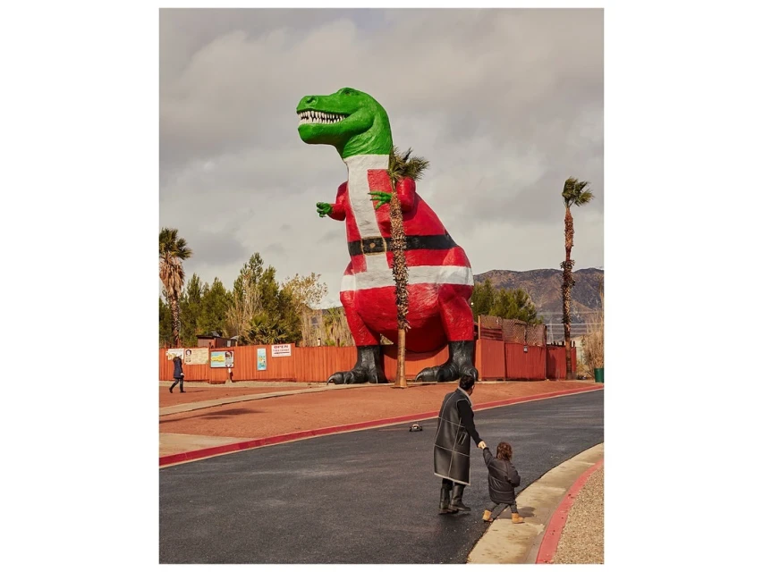 An adult and child walk towards a giant, green, Santa-suited T-Rex statue against a partly cloudy sky, creating a whimsical outdoor scene.