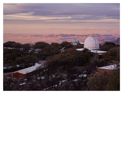 Observatory buildings with white domes sit amid trees under a sky with pastel clouds at sunset, viewed from an elevated position.