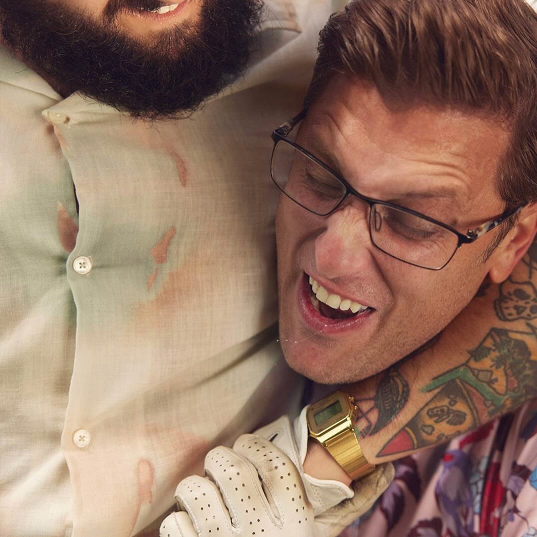 Two men in a close embrace, one with a beard and the other wearing glasses and a gold watch, captured in a humorous, indoor setting.