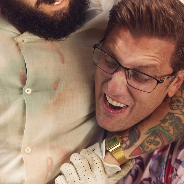 Two men in a close embrace, one with a beard and the other wearing glasses and a gold watch, captured in a humorous, indoor setting.