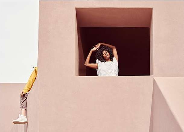 Woman in white top strikes a pose in a window while another person in yellow leans on a wall in a pink building, medium shot.