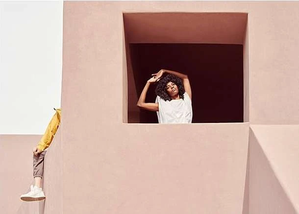 Woman in white top strikes a pose in a window while another person in yellow leans on a wall in a pink building, medium shot.