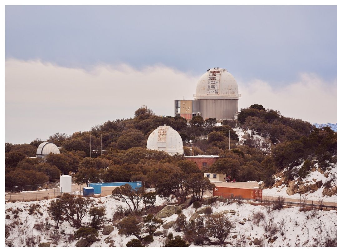 Wide shot of multiple white astronomical observatories atop a tree-covered hill in a winter landscape with a cloudy sky backdrop.