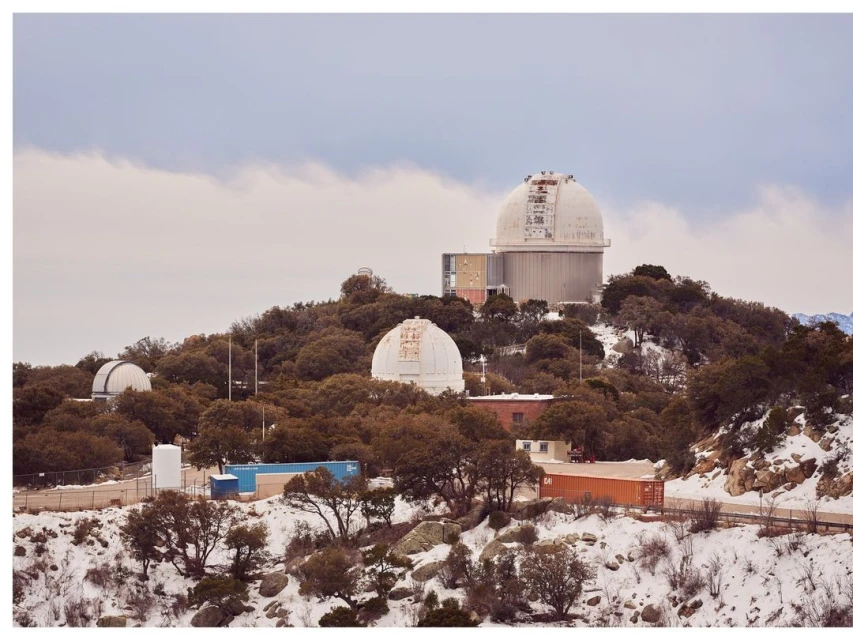 Wide shot of multiple white astronomical observatories atop a tree-covered hill in a winter landscape with a cloudy sky backdrop.
