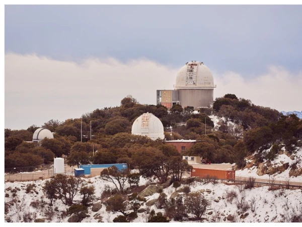 Wide shot of multiple white astronomical observatories atop a tree-covered hill in a winter landscape with a cloudy sky backdrop.