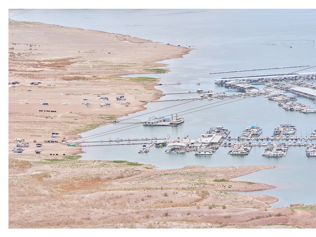 High-angle view of a marina on a partly dry lake, revealing arid shorelines, numerous boats docked, and vehicles near the shoreline.
