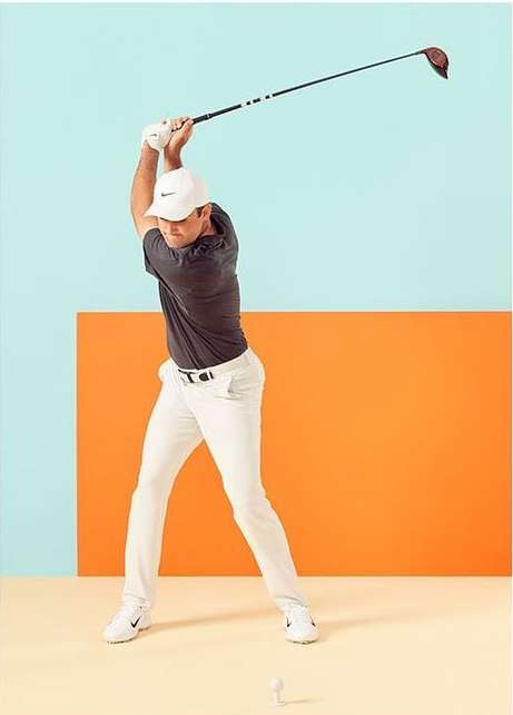 Male golfer in mid-swing wearing a white cap, grey shirt, and white pants, against a vibrant blue and orange background in a photo studio.