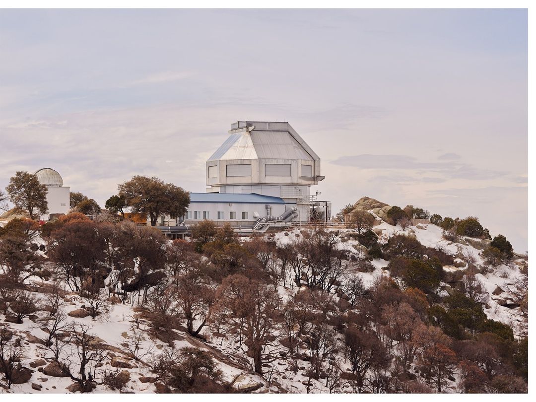 An observatory complex sits atop a snowy mountain covered in trees, under a pale sky. Clear atmosphere for astronomy research.