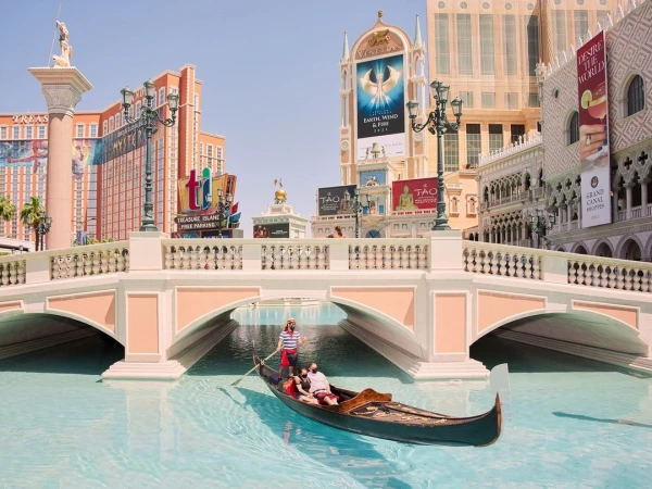 People ride a gondola on a canal with a gondolier in Las Vegas. The canal is under a bridge with hotels and casinos in the background.