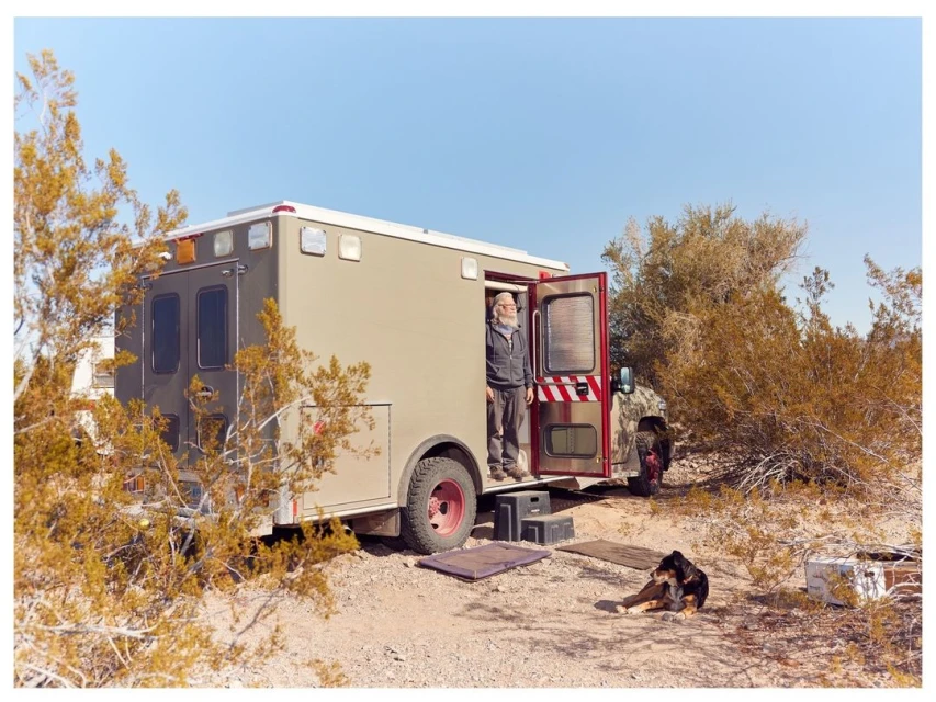 Ambulance converted into a camper in a desert landscape, with a senior man at the door and a resting dog on the ground.