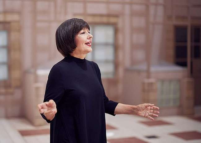 Candid portrait of a mature woman with short dark hair, wearing a black dress, and expressive hand gestures in an indoor setting.