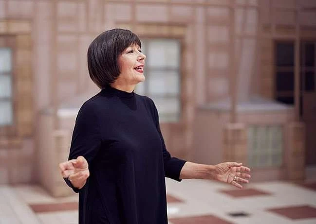Candid portrait of a mature woman with short dark hair, wearing a black dress, and expressive hand gestures in an indoor setting.