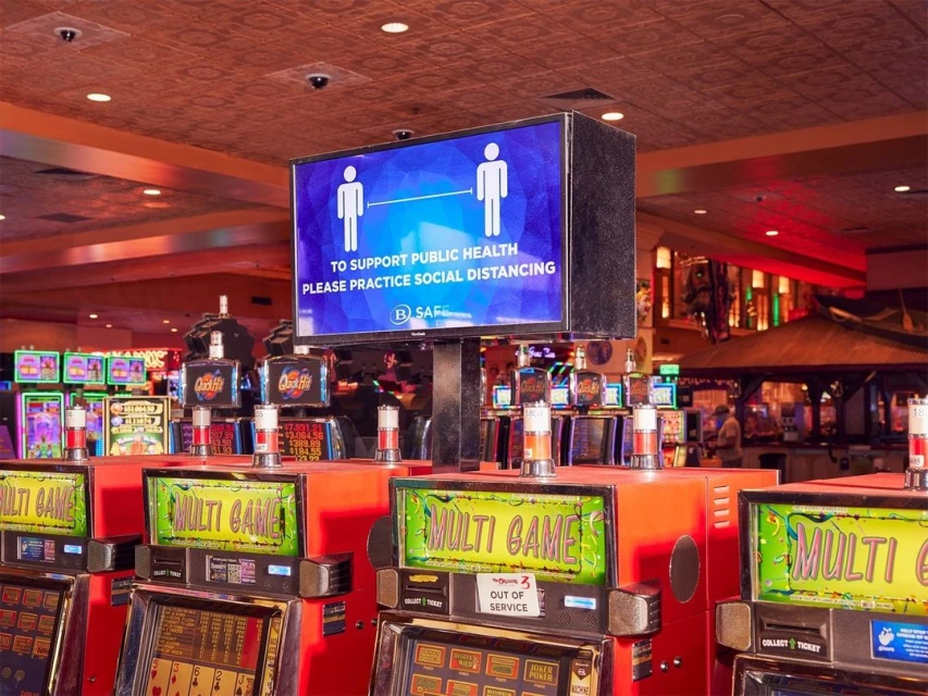 Indoor casino setting with rows of slot machines and a digital sign promoting social distancing for public health and safety.
