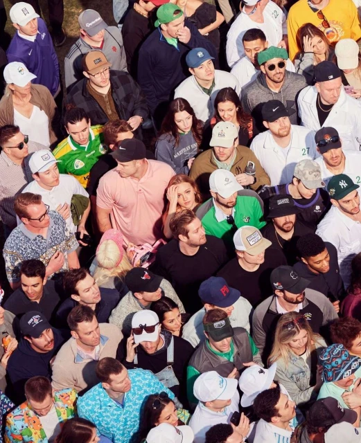 Aerial view of a large crowd of people gathered together, wearing various hats and clothing, suggesting a lively outdoor event or public gathering.