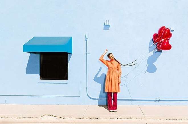 Woman in orange dress and pink pants stands next to a light-blue building, holding a bunch of red heart-shaped balloons.
